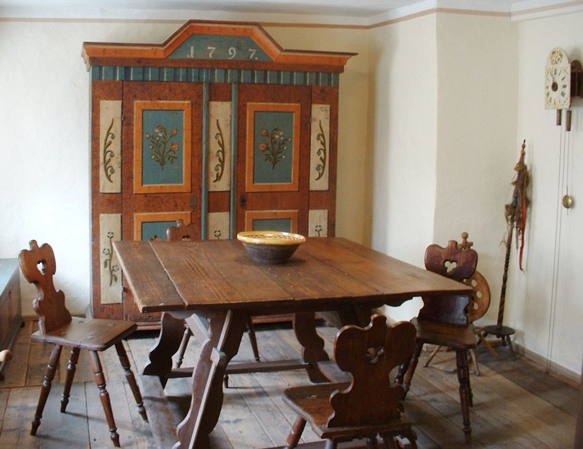 Blick in eine historisch anmutende Stube mit einem einfachen Esstisch und Stühlen aus Holz, einem großen bemalten Holzschrank, einem Spinnrad und einer Pendel-Wanduhr