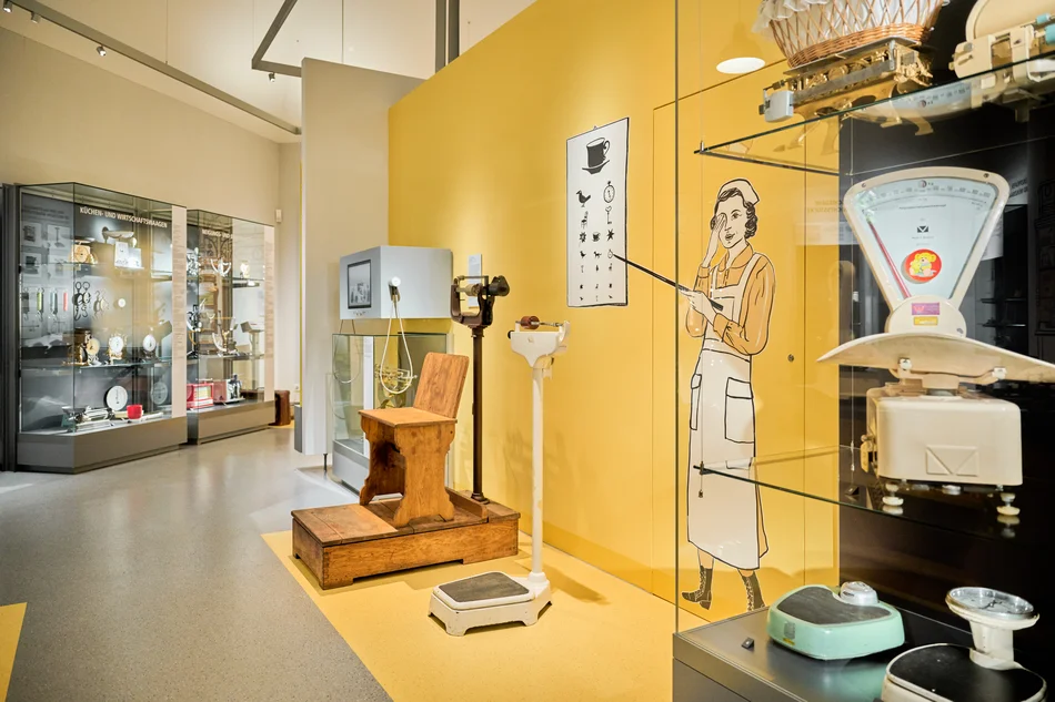Das Foto zeigt den Ausstellungsteil zu Waagen für den Gebrauch in Arztpraxen. Neben der Wandgrafik einer Krankenschwester stehen zwei historische Personenwaagen, in der benachbarten Vitrine ist eine alte Säuglingswaage zu sehen. Im Hintergrund ist eine Medienstation mit Bildschirm und Kopfhörer zu sehen.