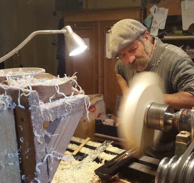 Ein Mann mit Schnauzbart und Baskenmütze bearbeitet an einer Drehbank ein Stück Holz. Späne fliegen.
