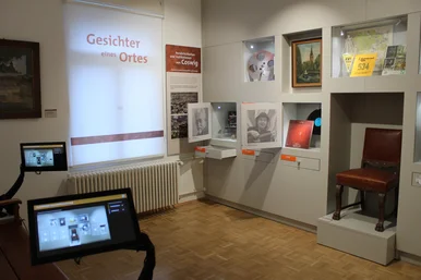 Ein kleiner Ausstellungsraum mit zwei Medienstationen, Wandvitrinen und Schubladen mit Ausstellungsexponaten. Auf der Fenster-Jalousie steht "Gesichter eines Ortes".