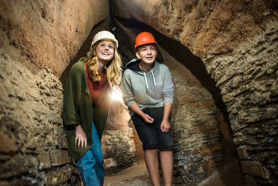Eine Frau und ein Jugendlicher mit Helm stehen in einem historischen Kellergang und schauen gespannt nach vorn.