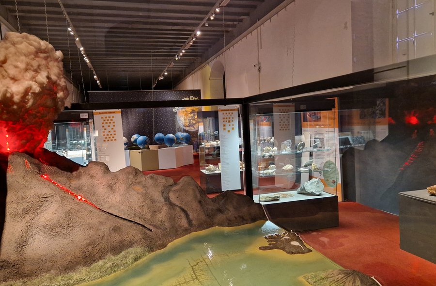 Der Blick in die Ausstellung umfasst mehrere große Vitrinen. Die Vitrine im Vordergrund enthält das Modell eines Vulkanausbruchs, im Hintergrund sind Vitrinen mit Planetenmodellen und Fossilien zu sehen.
