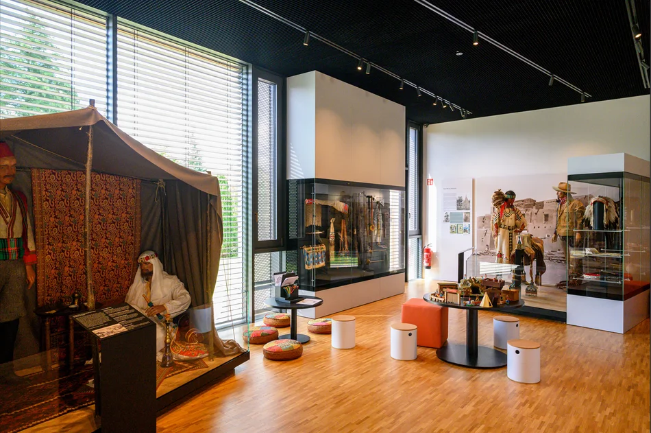 Ausstellungsbereich „Orient und Wilder Westen“ Das Foto zeigt einen Blick in den Dauerausstellungsbereich „Orient und Wilder Westen“. Im Vordergrund ist die Inszenierung einer lebensgroßen Figur in einem Orientzelt zu sehen, im Hintergrund der Bereich Indianistik mit zwei großen Vitrinen und zwei lebensgroßen Figuren vor einem großen Schwarzweißfoto einer nordamerikanischen Landschaft. Die beiden Bereiche trennt eine kleine Sitzgruppe mit Hockern, Sitzkissen und zwei runden Tischen.