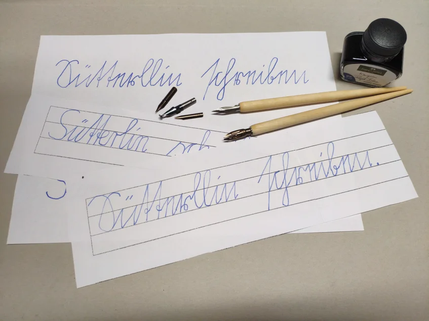 Sütterlin schreiben mit dem Federhalter Das Foto zeigt in Sütterlinschrift beschriebene Arbeitsblätter, ein Tintenfass und Schreibfedern.