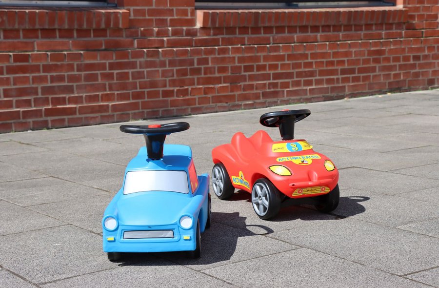 Auf einer betonierten Außenfläche des Industriemuseums steht neben einem typischen roten Bobbycar ein himmelblaues Bobbycar in der Form eines Pkw Trabant.