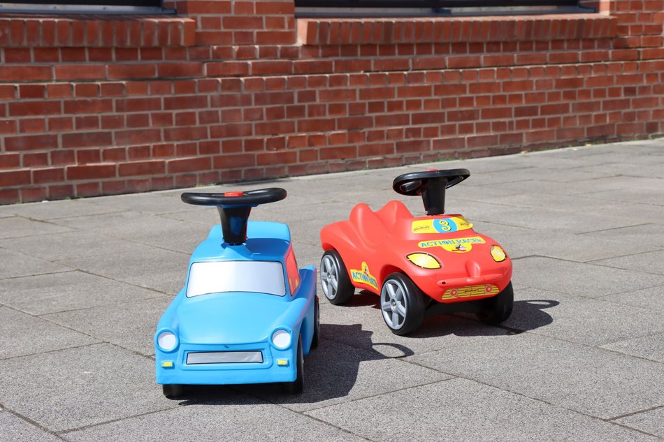 Auf einer betonierten Außenfläche des Industriemuseums steht neben einem typischen roten Bobbycar ein himmelblaues Bobbycar in der Form eines Pkw Trabant.