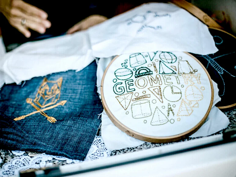 Links im Bild ein mit einem einfarbigen Ornament besticktes Stück Jeansstoff, rechts ein Stickrahmen mit handstickten geometrischen Formen und der gestickten Inschrift "Geometry"