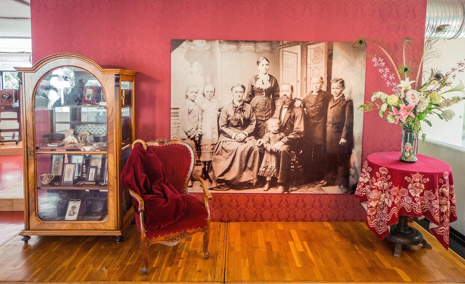 Auf einer roten Ausstellungswand hängt eine großformatige Reproduktion eines historischen, sepia-farbenen Familienfotos, dafür stehen ein historischer Sessel, ein runder Tisch mit elegantem Blumenbouquet und eine historische, mit diversen Gegenständen gefüllte Vitrine.