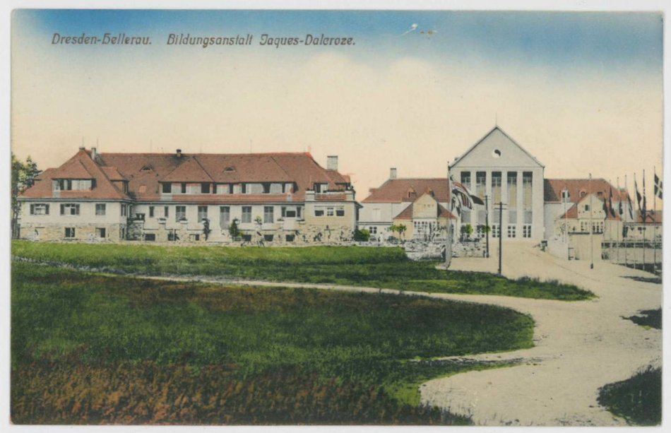 Das Foto zeigt eine kolorierte Postkarte aus der Zeit um 1912 mit der Aufschrift „Dresden-Hellerau. Bildungsanstalt Jacques-Daleroze.“  Das von Heinrich Tessenow im Stil der Reformarchitektur entworfene Gebäude beherbergt heute das Festspielhaus Hellerau.
