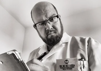 Ein Mann mittleren Alters mit Glatze, Bart, Brille, weißem Kittel und Klemmbrett blickt skeptisch in die Kamera.