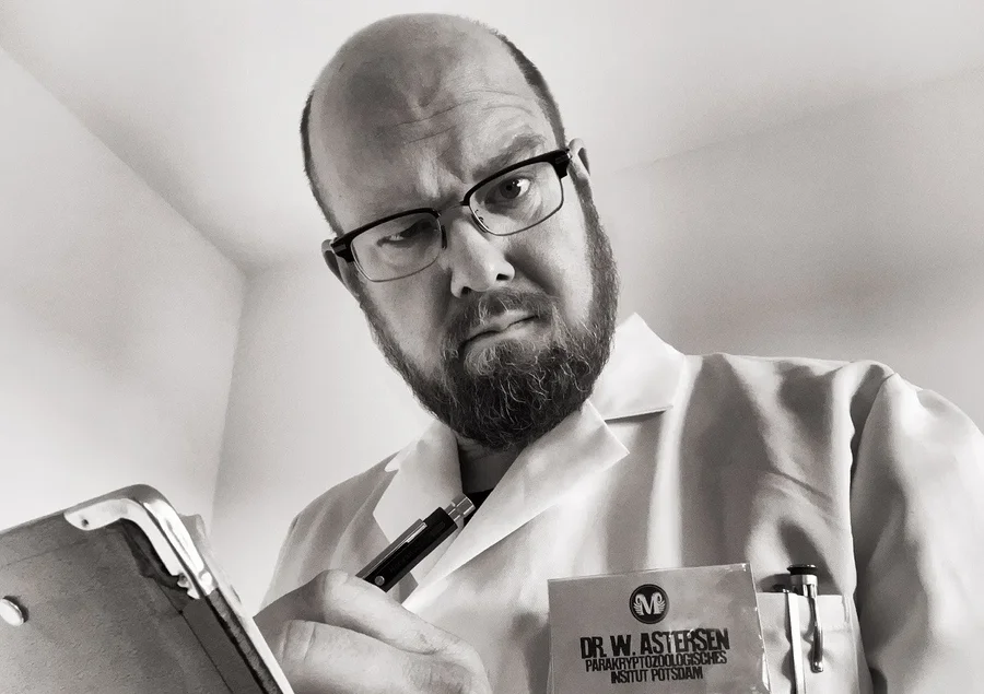 Ein Mann mittleren Alters mit Glatze, Bart, Brille, weißem Kittel und Klemmbrett blickt skeptisch in die Kamera.