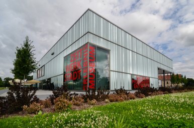 Eckansicht eines quaderförmigen modernen Funktionsgebäudes ragt vor einem bewölkten Himmel auf; an der dem Betrachter zugewandten Ecke befindet sich eine große Glasfläche, auf der in roten Großbuchstaben die Worte "ZeitWerkStadt" und "Erlebnismuseum" aufgebracht sind.