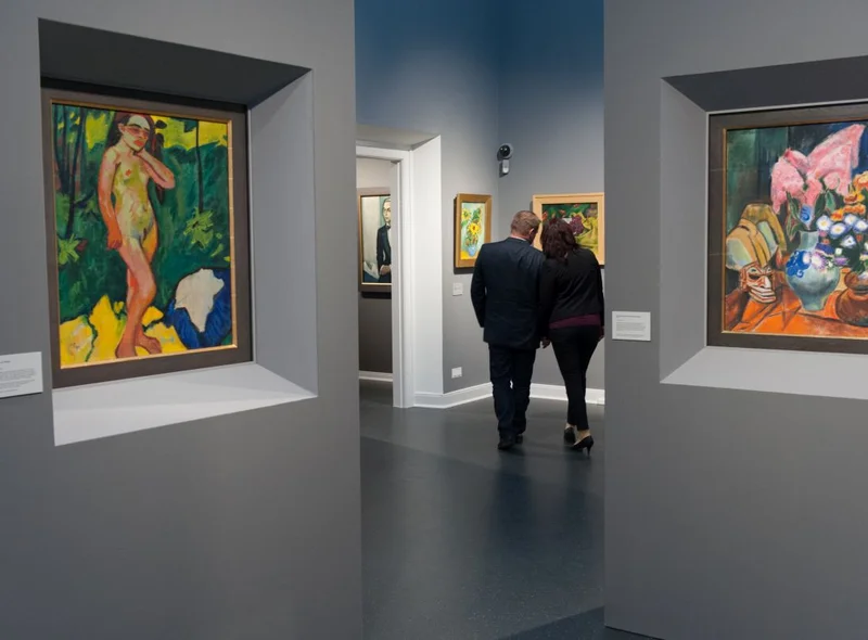 In einem Ausstellungssaal mit großformatigen Gemälden in kräftigen Farben stehen zwei Personen, die in die Kunstbetrachtung vertieft sind.