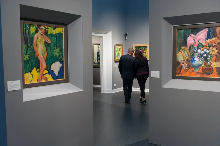 In einem Ausstellungssaal mit großformatigen Gemälden in kräftigen Farben stehen zwei Personen, die in die Kunstbetrachtung vertieft sind.