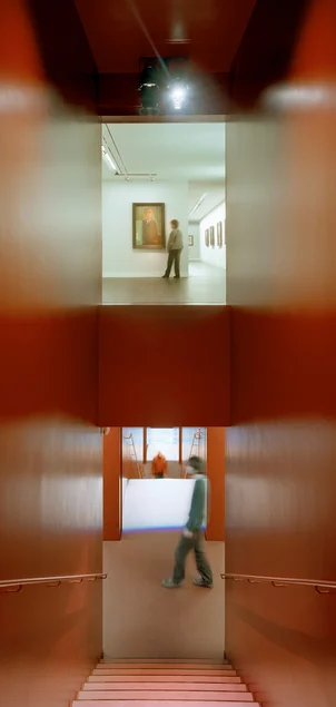 Das Foto zeigt einen Blick aus einem der Obergeschosse des Museum Gunzenhauser in das schmale, steile, ganz in dunklem Rot gehaltene Treppenhaus. Geradeaus kann man durch ein großes Fenster in den gegenüberliegenden Ausstellungsraum auf derselben Etage schauen, nach unten sieht man bis zum nächsten Treppenabsatz, den eine Person quert, sowie weiter nach unten bis zum übernächsten Treppenabsatz.