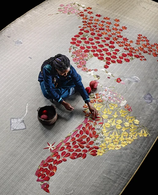 Eine Person webt einen großen Teppich mit Blumen, welche die Form von Südamerika bilden