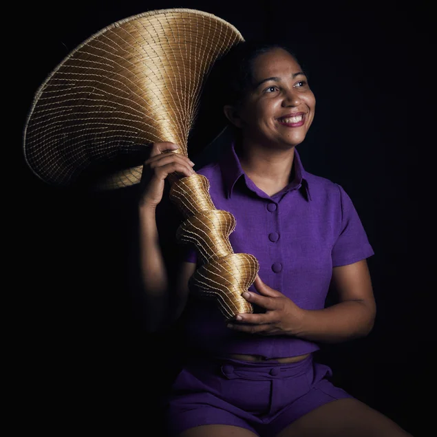 Eine Person hält lächelnd ein großes, trichterförmiges, golden schimmerndes Objekt aus Gras in den Händen.