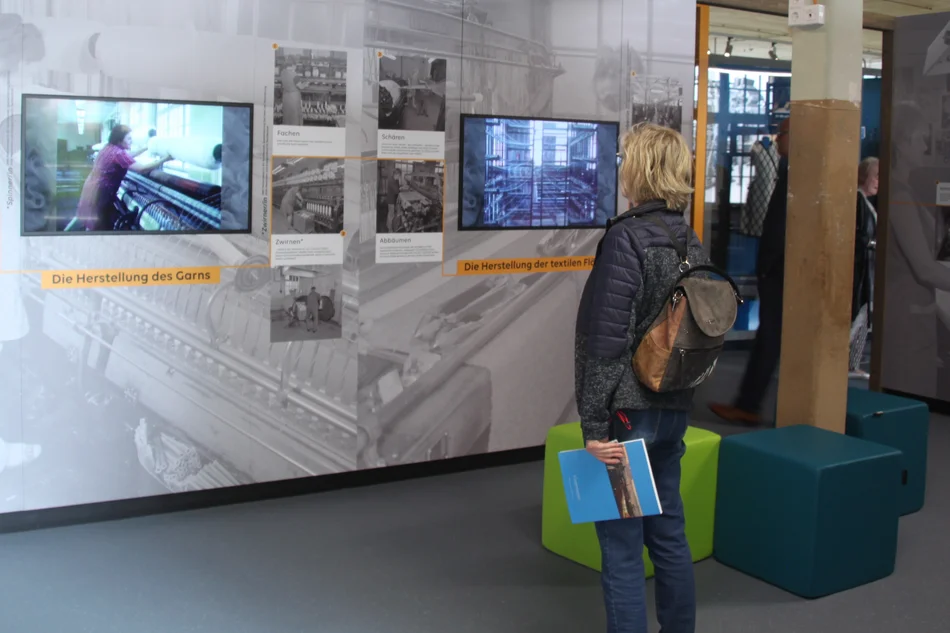 Eine Frau steht vor einer mit Bildern und Texten sowie zwei Monitoren ausgestatteten Ausstellungswand.