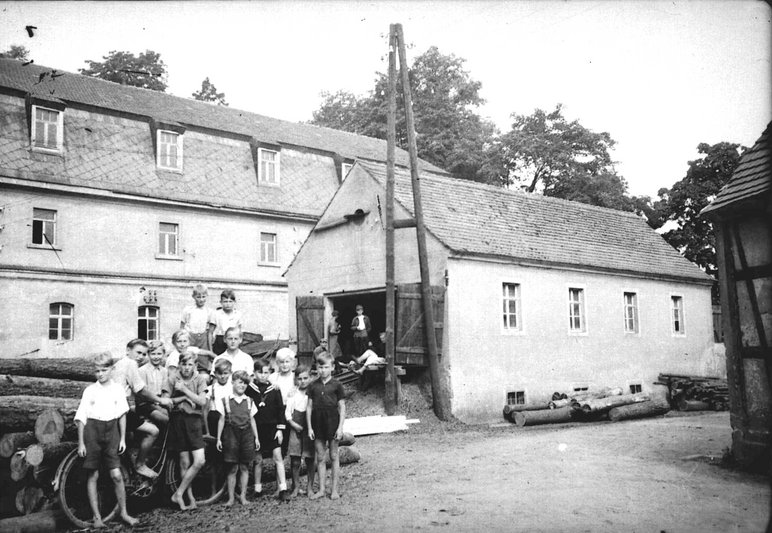 Historisches Schwarzweißfoto, auf dem eine Gruppe Kinder vor einem gewerblich genutzten und einem vermutlich zum Wohnen genutzten Gebäude steht