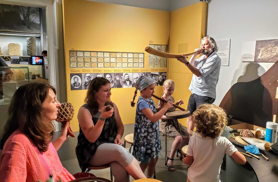 In einem Ausstellungsraum probieren sich Kinder und Erwachsene an verschiedenen Musikinstrumenten aus. Ein Mann bläst in ein langes, geschwungenes Horn.