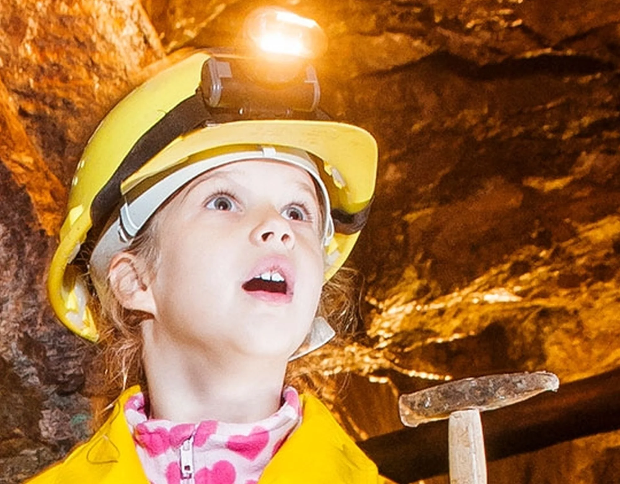 Kind im Bergwerk Das Foto zeigt ein staunendes Kind im Bergwerk, es trägt einen gelben Helm mit Grubenlampe und einen gelben Mantel und hält einen Hammer in der Hand.