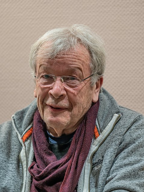 Ein älterer Mann mit grauen Haaren, rahmenloser Brille, grauer Jacke und weinrotem Schal blickt freundlich in die Kamera.
