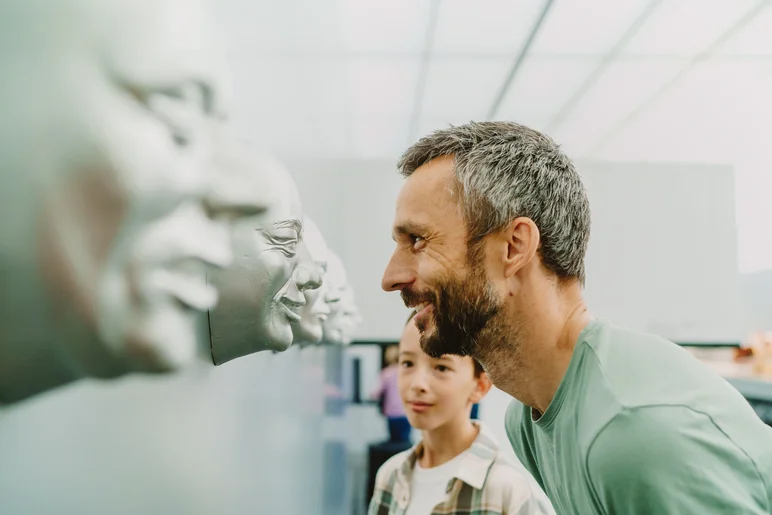 In einem Ausstellungsraum stehen ein Mann und ein Junge Auge in Auge mit einer Reihe von Gipsabdrücken menschlicher Gesichter