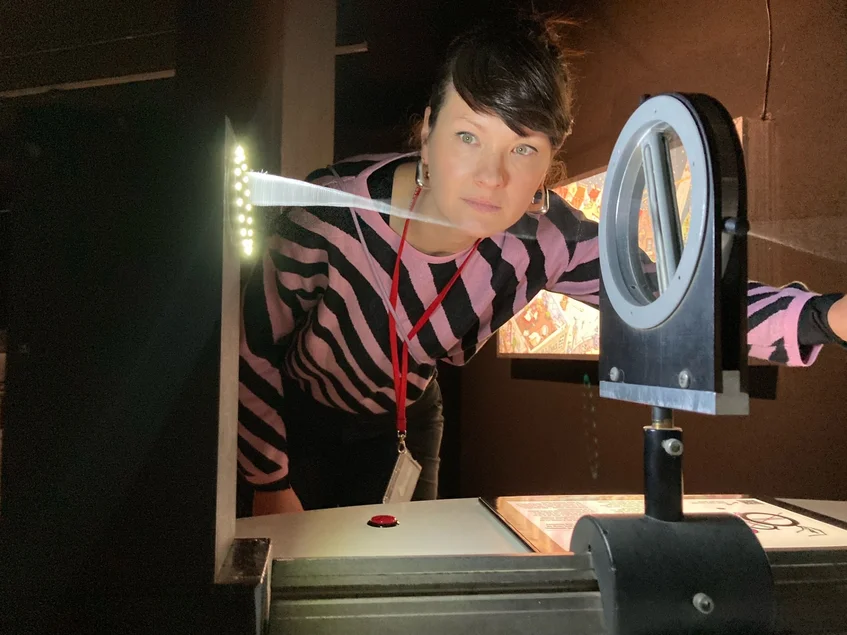 Ausstellungsbesucherin führt an opto-akustischen Instrumenten ein Experiment durch. Links ist eine Lichtquelle erkennbar.