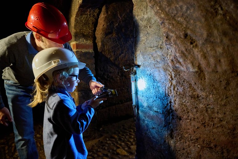 Ein Kind mit Helm leuchtet mit einer Taschenlampe Gestein unter dem Schloss an, hinter ihm ein Erwachsener mit Helm.