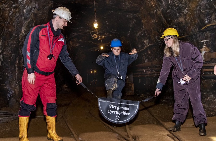 Erlebnisführung: „Bergmann ehrenhalber“ In einem Stollen stehen ein Mitarbeiter der Zinngrube in Schutzkleidung (schwarz-rote Jacke und Hose, gelbe Gummistiefel und weißer Schutzhelm) und eine Besucherin (erwachsene Frau) in Schutzkleidung (brauner Overall, gelber Schutzhelm, schwarze Gummistiefel). Beide halten einen schwarzen Lederriemen fest, an dem in der Mitte ein halbrundes Lederstück angebracht ist. Auf diesem steht "Bergmann ehrenhalber" und darüber gekreuzt Schlägel und Eisen in weißer Schrift. Über diesen Lederriemen springt ein zweiter Besucher (Kind) in Schutzkleidung (blauer Overall und Schutzhelm, schwarze Gummistiefel).