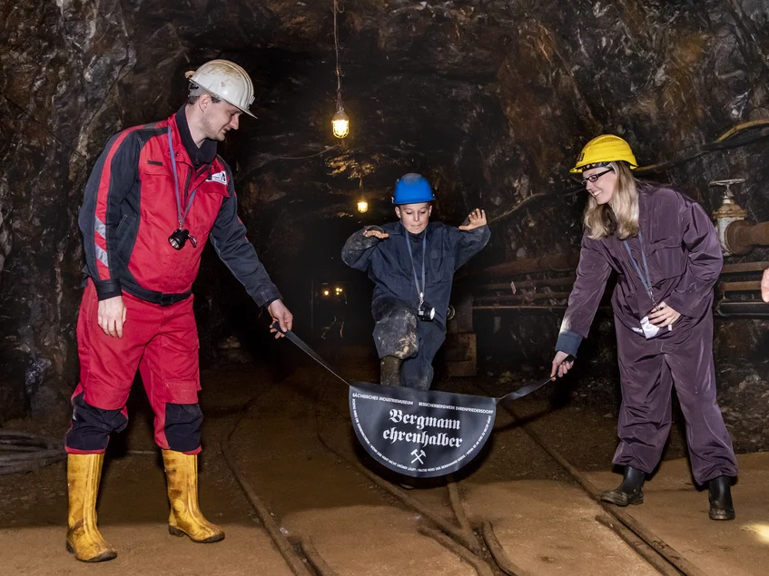 Erlebnisführung: „Bergmann ehrenhalber“ In einem Stollen stehen ein Mitarbeiter der Zinngrube in Schutzkleidung (schwarz-rote Jacke und Hose, gelbe Gummistiefel und weißer Schutzhelm) und eine Besucherin (erwachsene Frau) in Schutzkleidung (brauner Overall, gelber Schutzhelm, schwarze Gummistiefel). Beide halten einen schwarzen Lederriemen fest, an dem in der Mitte ein halbrundes Lederstück angebracht ist. Auf diesem steht "Bergmann ehrenhalber" und darüber gekreuzt Schlägel und Eisen in weißer Schrift. Über diesen Lederriemen springt ein zweiter Besucher (Kind) in Schutzkleidung (blauer Overall und Schutzhelm, schwarze Gummistiefel).