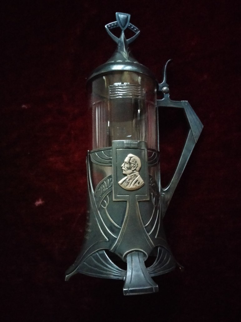 In typischer Jugendstil-Manier gestaltetes Metallobjekt mit Griff und Deckel, das als Glashalter dient