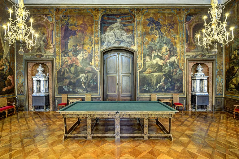 Ein prunkvoll ausgestatteter Saal mit einem Billiardtisch im Zentrum und reich verzierten Goldledertapeten im Hintergrund