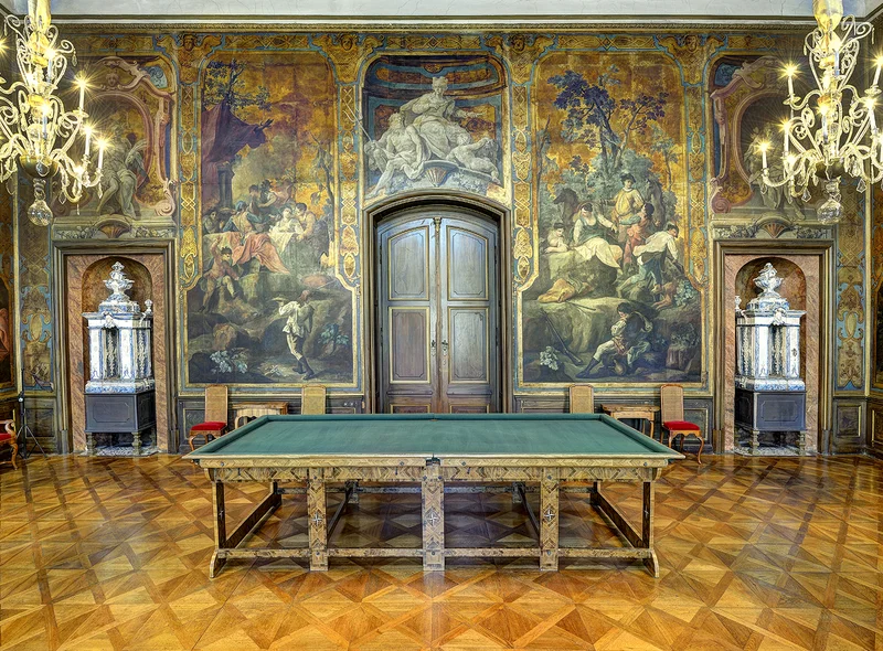 Ein prunkvoll ausgestatteter Saal mit einem Billiardtisch im Zentrum und reich verzierten Goldledertapeten im Hintergrund