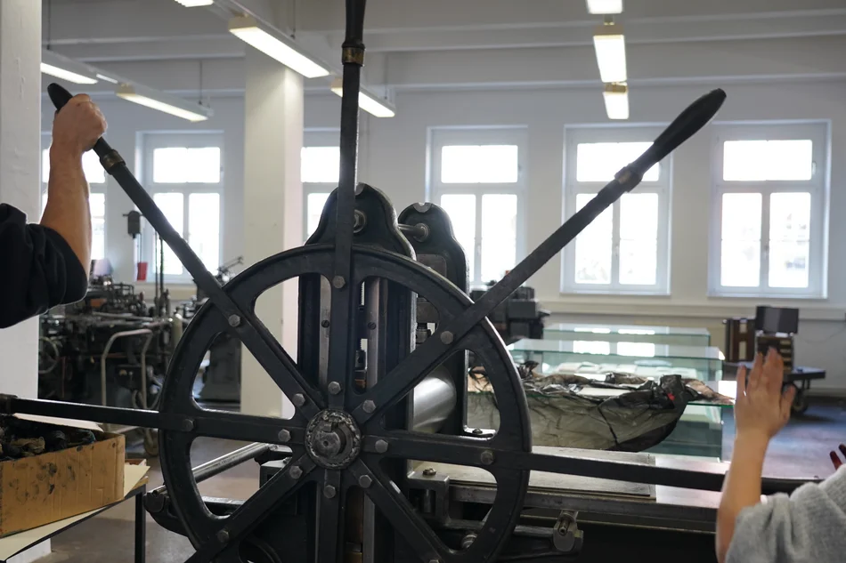 In einem großen Drucksaal dreht eine Hand am Rad einer historischen manuellen Druckerpresse.