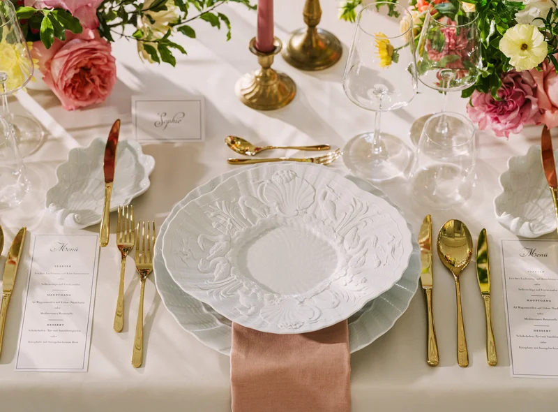 Detailaufnahme einer festlich gedeckten Tafel mit weißem Meißener Porzellan und goldfarbenem Besteck, rings herum Blumen