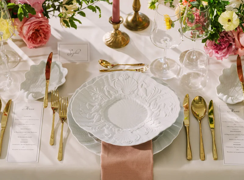 Detailaufnahme einer festlich gedeckten Tafel mit weißem Meißener Porzellan und goldfarbenem Besteck, rings herum Blumen