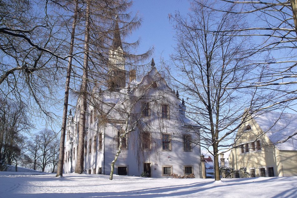 Das Foto zeigt eine winterliche Außenansicht von Schloss Burgk in Freital. Boden und Dächer sind schneebedeckt, das Schloss ist halb verdeckt durch einige kahle Bäume, der Himmel ist wolkenlos und strahlend blau.