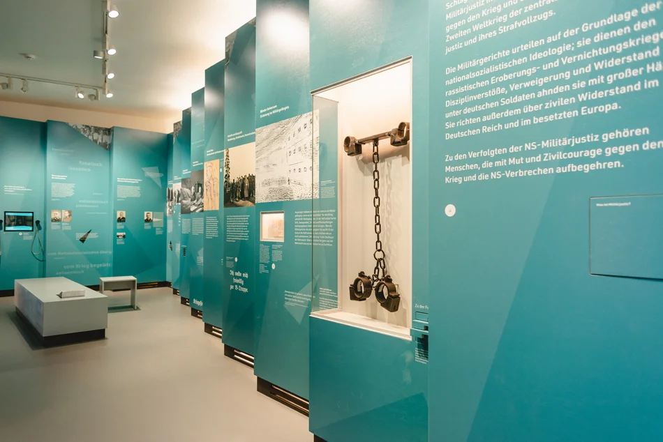 Blick in einen Ausstellungsraum, an dessen Wände in Türkis gehaltene Ausstellungsmodule angeordnet sind. Diese enthalten sowohl Texte und Fotos als auch Medienstationen und Wandvitrinen.