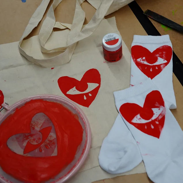 Logo-Motiv ist ein mit einem Auge versehenes rotes Herz. Dieses ist auf einen Stoffbeutel wie auch auf ein Paar weiße Socken gedruckt.
