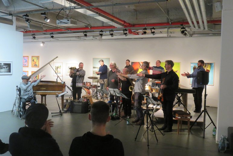 Zehn Musiker spielen verschiedene Instrumente in einem Kunstausstellungsraum, hinter ihnen hängen diverse Gemälde.
