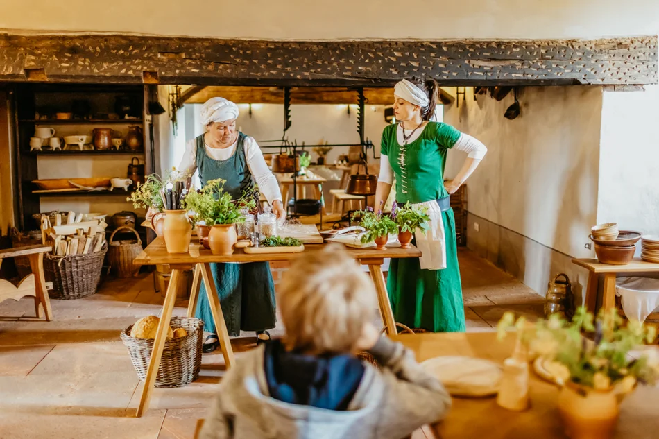 Zwei Küchenmägde zeigen verschiedene Getreidesorten und Gartenkräuter in einer historischen Küche.