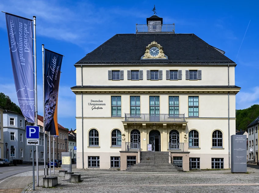Deutsches Uhrenmuseum Glashütte Ein alleinstehendes, vierstöckiges historisches Gebäude mit Portal. Auf dem Platz davor wehen zwei Fahnen des Uhrenmuseums.