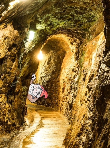 Zwergenkönig Foto eines beleuchteten Bergwerkstollens, in das eine cartoonhafte Zeichnung eines frechen Zwerges eingefügt ist