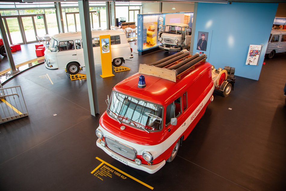 Blick von der Empore auf drei im Erdgeschoss ausgestellte Transporter des Typs "Barkas" – zwei davon weiß lackiert, der dritte, im Vordergrund befindliche als Feuerwehrauto ausgestattet und rot lackiert