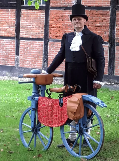 Ein Mann in Frack und Zylinder im Stil des 19. Jahrhunderts steht hinter einem historischen Holz-Fahrrad.