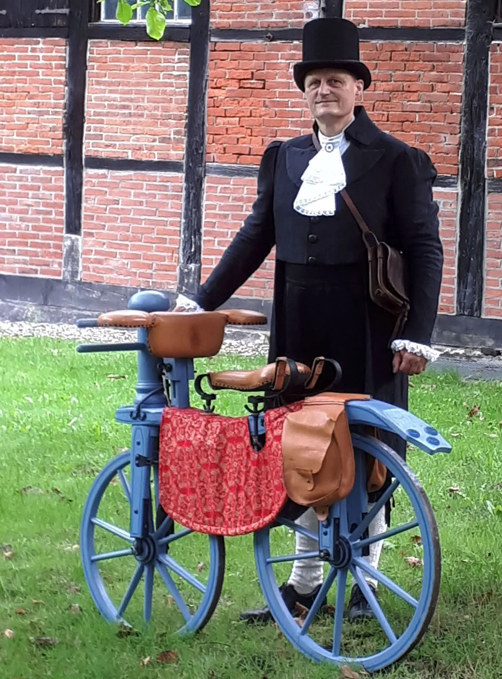 Ein Mann in Frack und Zylinder im Stil des 19. Jahrhunderts steht hinter einem historischen Holz-Fahrrad.