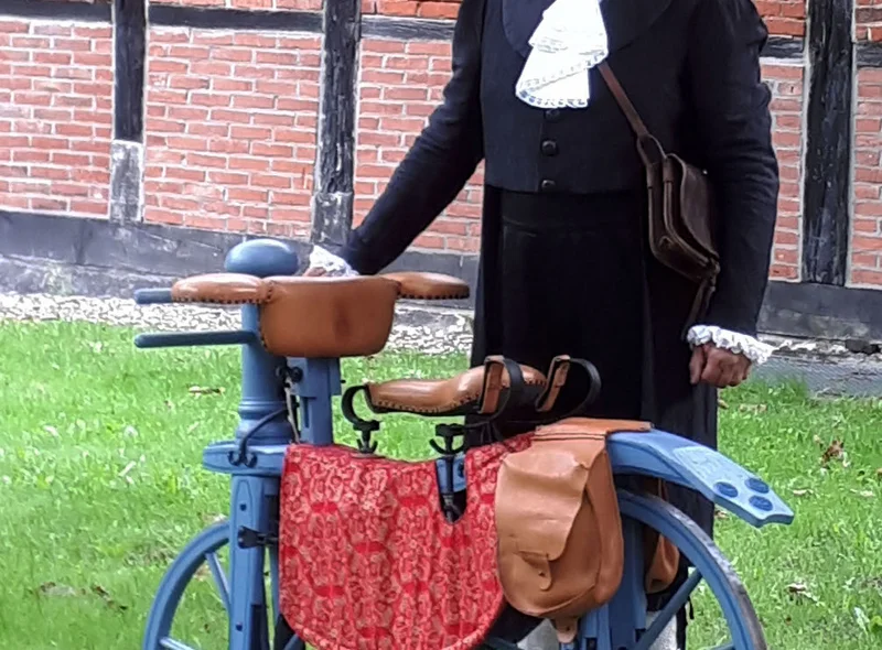 Ein Mann in Frack und Zylinder im Stil des 19. Jahrhunderts steht hinter einem historischen Holz-Fahrrad.