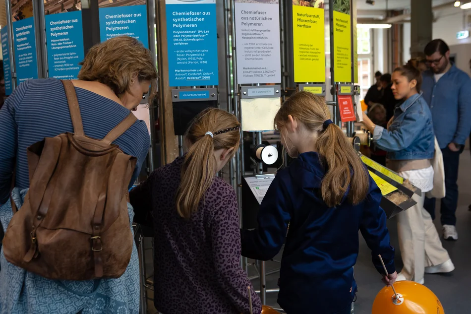 Turm der Tuche An einem kreisförmig angeordneten Ausstellungselement, an dem Text- und Bildtafeln angebracht sind, stehen mehrere Besucherinnen und Besucher.