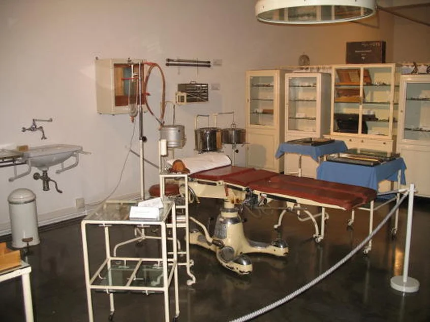 In einem klinisch anmutenden Raum steht mittig ein historischer OP-Tisch, um ihn herum diverse alte medizinische Geräte.